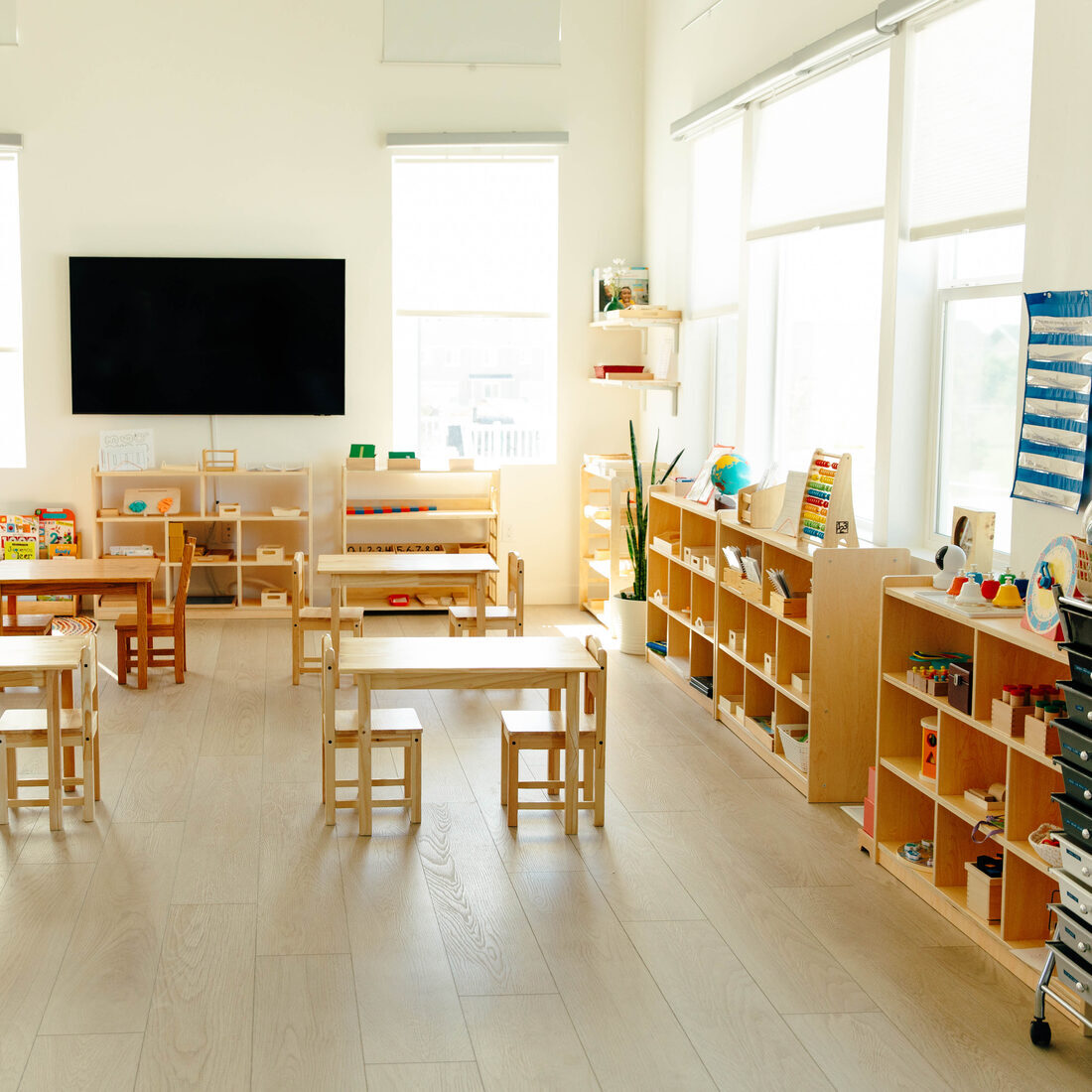 The main classroom: child-height tables and chairs, low shelving along one wall, tall windows on two sides.