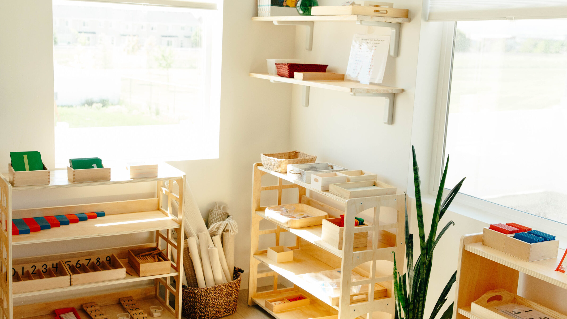 The classroom on a bright morning: low wooden shelves of Montessori materials, a basket of rolled work mats, and daylight through two windows.