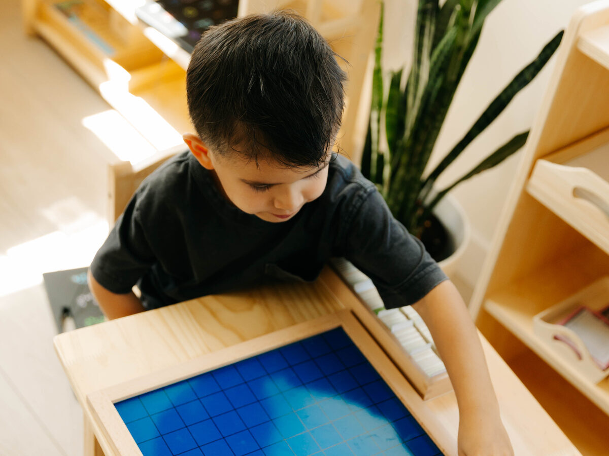 A preschooler working alone at a table, placing a numbered tile on the blue hundred board.